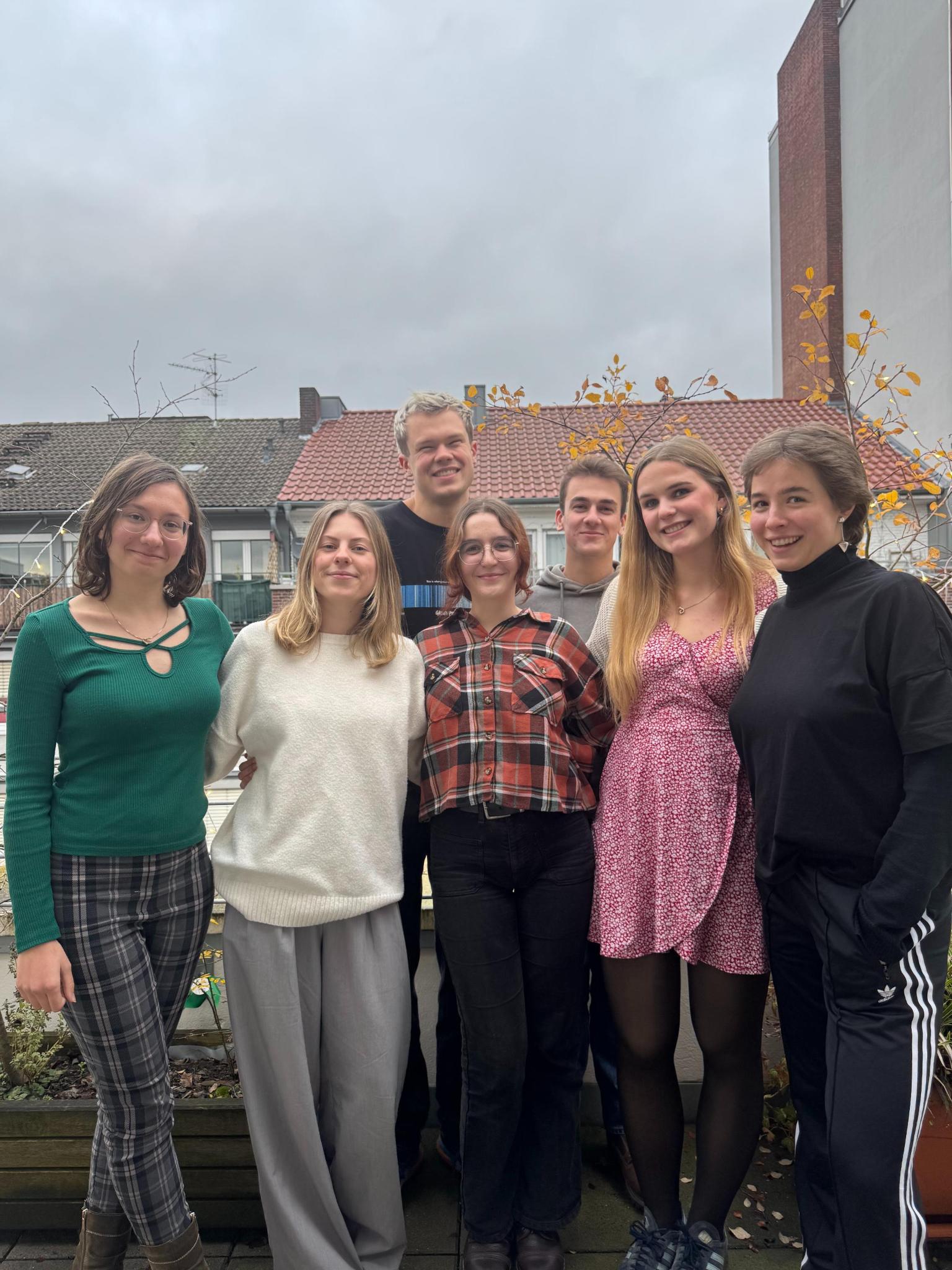 Die Mitglieder des Vorstandes stehen in zwei Reihen auf einem Balkon und lächeln in die Kamera. Hinter ihnen sind Blumentöpfe mit Pflanzen und ein anderes Haus zu sehen.